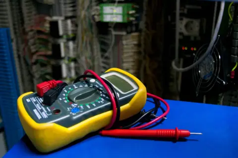 Żółty multimetr z czerwonymi i czarnymi przewodami na tle szafy elektrycznej.