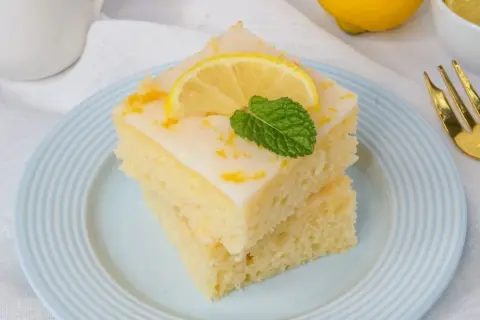 Zwei Stücke zitronenkuchen mit joghurt, garniert mit Zitronenscheibe und Minze, auf blau-weiß gestreiftem Teller.