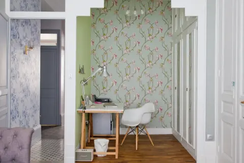 Un pan de mur de salon est habillé d'un papier peint floral aux fleurs roses sur fond vert pâle. Un bureau et une chaise blanche complètent l'espace.