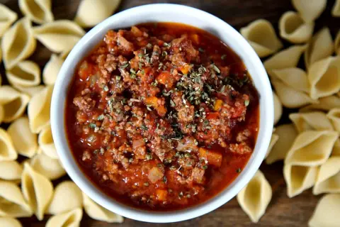 Herzhaftes Bolognese-Rezept mit Hackfleisch und Tomaten, garniert mit Kräutern, umgeben von ungekochten Nudeln.