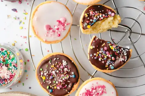 Bunte mini Amerikaner mit Schokoladen- und Zuckerguss, bestreut mit Streuseln und Herzen, auf einem Kuchengitter.