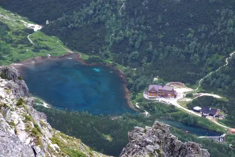 Widok na zielony staw kieżmarski otoczony lasem i górami. W pobliżu schronisko i ścieżki.
