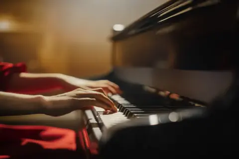 Dłonie kobiety w czerwonej sukni grają na fortepianie. Proces budowy fortepianu jest fascynujący, a dźwięk instrumentu porusza serca.