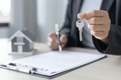 Persona firma un contrato de alquiler con opción a compra, sosteniendo la llave de una casa en miniatura.