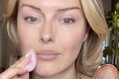 Eine Frau trägt mit einem Schwämmchen puder auf ihr Gesicht auf. Ihr blondes Haar umrahmt ihr Gesicht, während sie sich auf ihr Make-up konzentriert.
