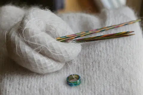 Ein Knäuel weiches, flauschiges Mohairgarn liegt neben bunten Stricknadeln auf einem Strickstück. Was ist Mohair? Es ist eine luxuriöse Faser.