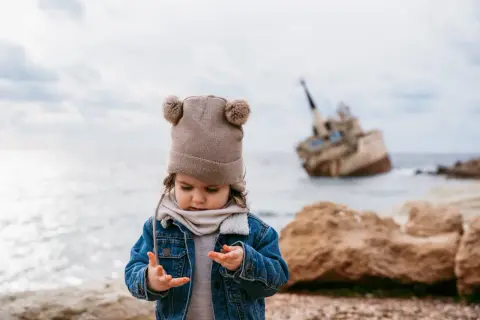 Małe dziecko w czapce z pomponami bawi się na plaży na Cyprze, w tle wrak statku.