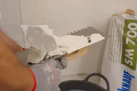 Man applying morteros para fachadas con llana dentada. Bolsa de producto de construcción al fondo.