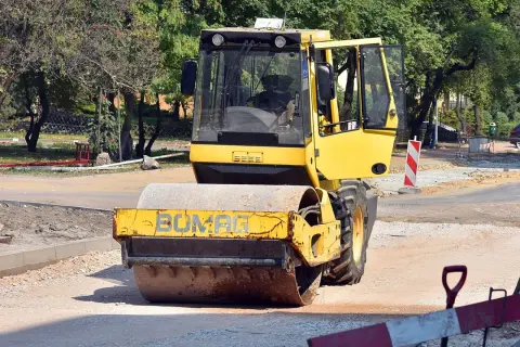 Żółty pojazd wolnobieżny, walec drogowy Bomag, pracuje na budowie drogi, ubijając nawierzchnię.