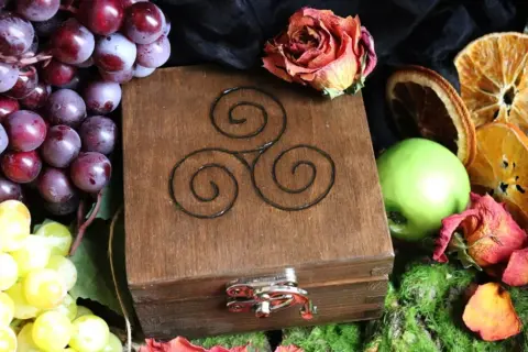 Drewniana szkatułka z symbolem triskelion, otoczona winogronami, suszonymi pomarańczami, jabłkiem i różą.
