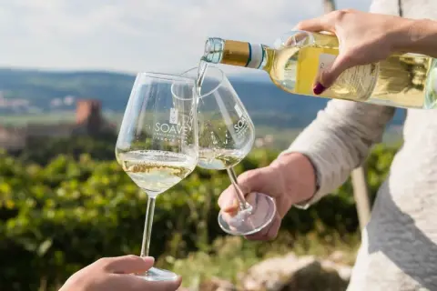 Versando vino bianco in calici con logo Soave, sullo sfondo un paesaggio collinare con vigneti e un castello. Un brindisi al Soave Superiore DOCG.