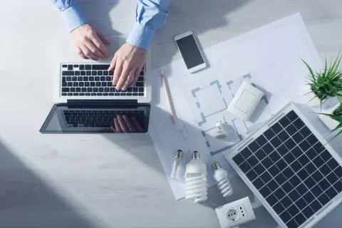 Analiza sprawności paneli fotowoltaicznych na laptopie. Na biurku projekt, żarówki, gniazdko i telefon.