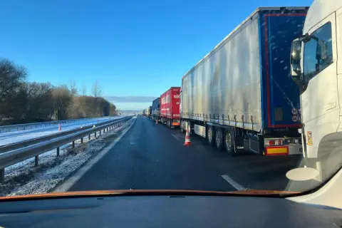 Longue file de véhicules poids lourds à l'arrêt sur une autoroute enneigée sous un ciel bleu clair.
