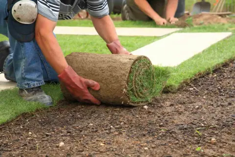 Mann mit Handschuhen rollt Rasen aus, um ihn zu verlegen. Ein weiterer Mann im Hintergrund hilft.