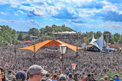 Tausende feiern auf dem Fusion Festival Ort unter blauem Himmel. Bunte Zelte und Bühnen prägen die Szenerie.