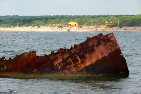 Rdza rdzy, wrak statku na Bałtyku. Czy to symbol, dlaczego wody bałtyku są znacznie zanieczyszczone? W tle plaża z ludźmi.
