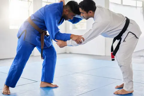 Dwóch mężczyzn w kimonach do BJJ, jeden w niebieskim z brązowym pasem, drugi w białym z czarnym pasem, ćwiczy chwyty na macie.
