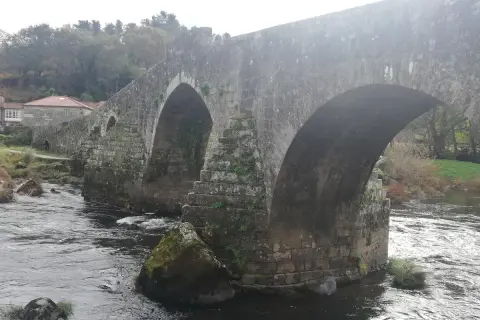 El histórico puente Maceira cruza un río caudaloso, con casas de tejados rojos y árboles frondosos al fondo.