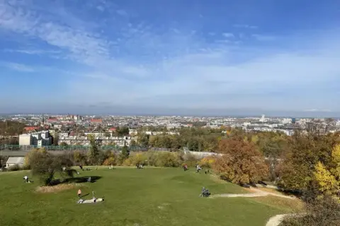 Widok na Kraków z zielonego wzgórza, gdzie ludzie odpoczywają. W oddali rozciąga się panorama miasta, a jesienne drzewa dodają uroku. To idealne miejsce na spacer, jak na **Kopcu Kraka**.