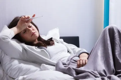 Kobieta leży w łóżku z termometrem na czole, czując się źle. Może potrzebuje nalgesin na ból.