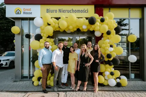 Grupa ludzi pozuje przed biurem nieruchomości ozdobionym żółtymi balonami.