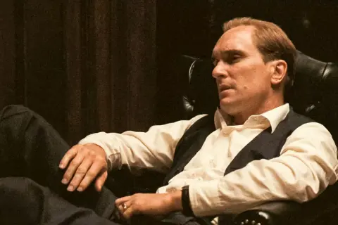 Robert Duvall jako Tom Hagen w fotelu, zamyślony, w białej koszuli i kamizelce.
