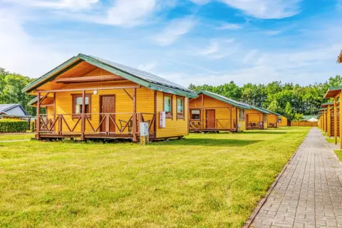 Drewniane domki letniskowe w ośrodku Błękitna Laguna Karwia, z werandami i panelami słonecznymi na dachach, stoją na zielonej trawie.