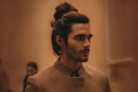 Un homme au chignon bas homme élégant, portant une veste texturée, pose avec assurance.