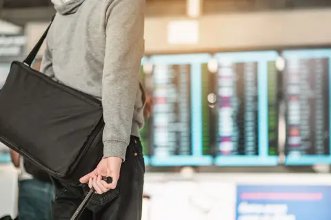 Mann mit schwarzer Reisetasche und Koffer vor Anzeigetafel am Flughafen.