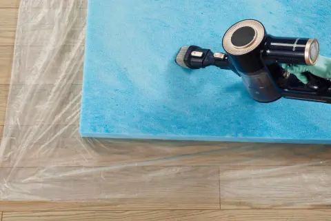 A gloved hand uses a vacuum cleaner with a brush attachment to clean a blue foam mattress topper laid on plastic sheeting.