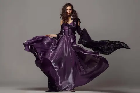 A model in a flowing purple gown and black lace sleeves poses for studio fashion photography.