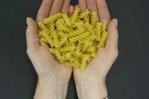 Dłonie trzymające porcję żółtego makaronu fusilli na ciemnym tle.