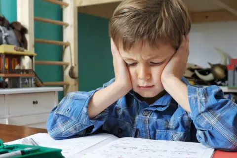 Chłopiec z problemem z koncentracją u dziecka, z rękami na uszach, siedzi przy biurku, próbując odrobić lekcje.