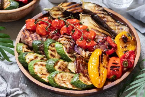 Una colorida parrillada de verduras con calabacín, berenjena, tomates cherry, pimientos y cebolla, servida en una bandeja de madera.