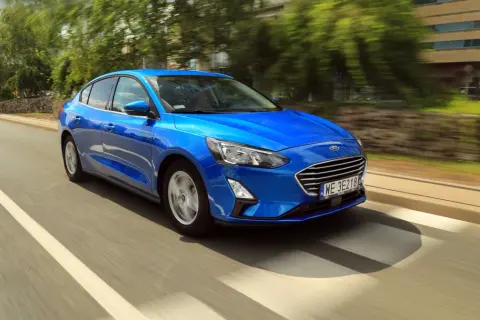 Błękitny Ford Focus sedan pędzi po miejskiej drodze, mijając drzewa i budynki.