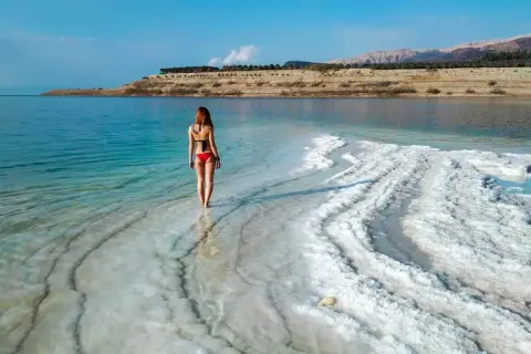 Kobieta w czerwonym bikini spaceruje po krystalicznej plaży. To właśnie Morze Martwe, gdzie można unosić się na wodzie.
