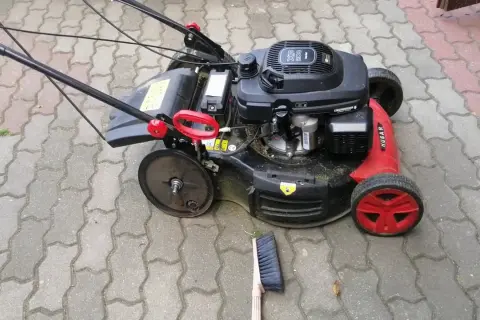 Kosiarka z czerwonymi detalami i solidnymi kółkami do kosiarki, gotowa do pracy na podjeździe z kostki brukowej. Obok leży miotka.