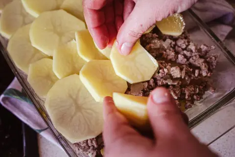 Dłonie układają plastry ziemniaków na mielonym mięsie, tworząc prostą zapiekankę ziemniaczaną.