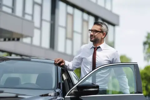 Biznesmen w białej koszuli i krawacie opiera się o drzwi czarnego samochodu. Czeka na niego kolejna kilometrówka.