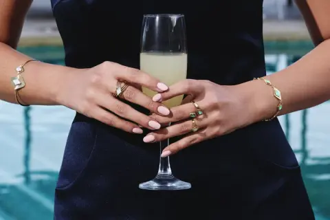 Manos con uñas rosadas y varios anillos dorados, sosteniendo una copa de champán. Brazaletes dorados adornan ambas muñecas.