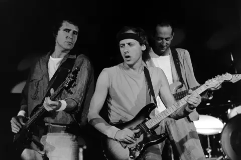 Trzech członków Dire Straits gra na gitarach podczas koncertu.
