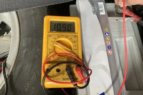Multímetro amarillo muestra 10.90V al comprobar bateria coche. Se ve la batería, un neumático y cables.