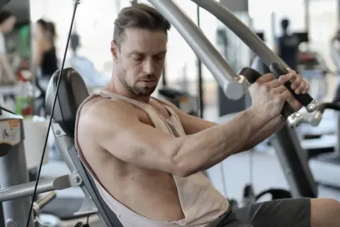 Mann trainiert im Fitnessstudio, um schnell zuzunehmen. Er sitzt auf einem Gerät und drückt die Griffe nach vorne.