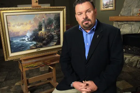 A man in a suit sits beside a Thomas Kinkade painting of a lighthouse on a cliff.