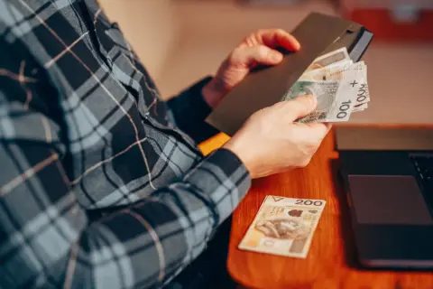 Osoba w kratę liczy pieniądze, przygotowując donos do urzędu skarbowego. Obok leży laptop i banknot 200 zł.