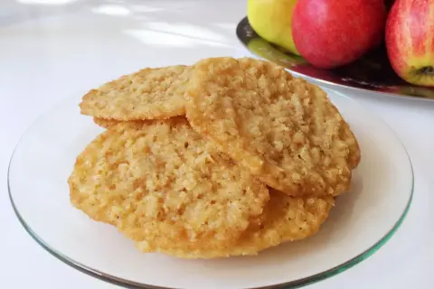 Tre gyllene glutenfria havreflarn ligger staplade på en glasfat. I bakgrunden syns äpplen och päron.