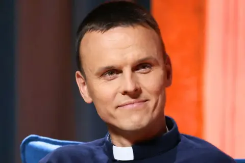 Uśmiechnięty ksiądz z osiedla, młody duchowny w sutannie, z uwagą słucha rozmówcy.