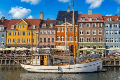 Ein weißes Segelschiff liegt im Hafen vor bunten Häusern. Menschen sitzen in Cafés am Wasser. Ein perfekter Tag im kopenhagen sommer.