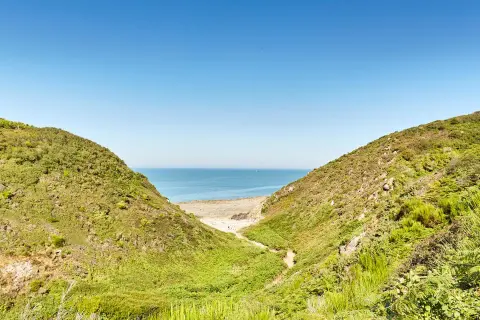 Vue sur la mer à travers la vallée du Lude, bordée de collines verdoyantes et d'une plage de sable.