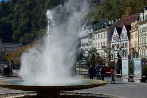 Der sprühende geysir karlsbad mit dampfender Fontäne vor bunten Häusern und grünen Bäumen.